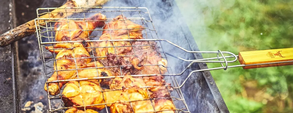 Chicken pieces cooking in a wire grilling basket over a smoky outdoor grill.