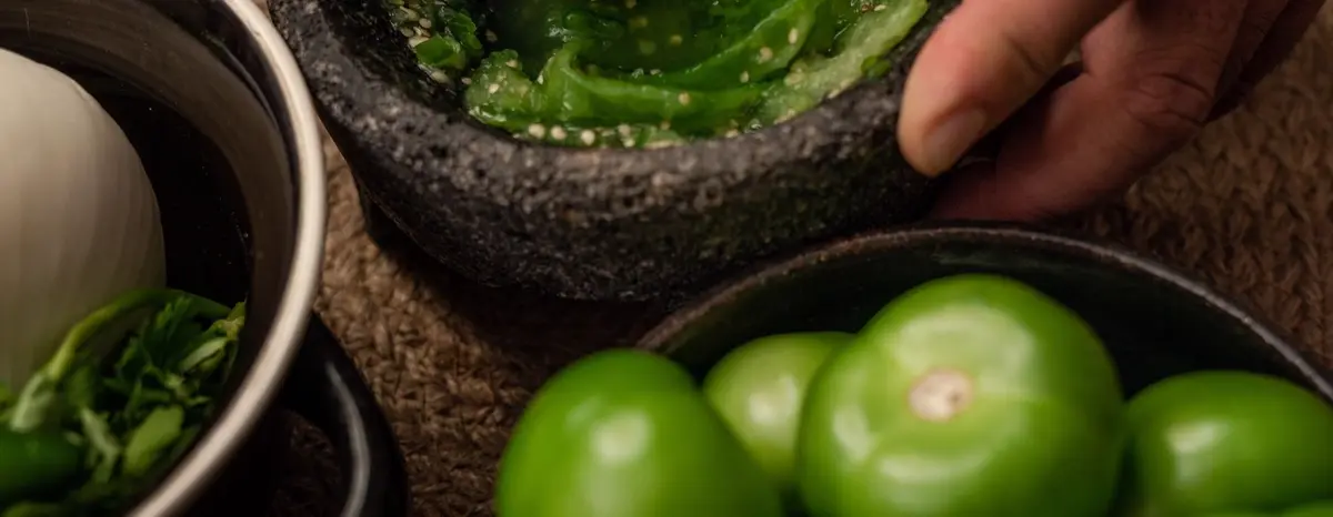 Green salsa being prepared in a stone mortar beside whole green tomatoes.
